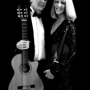 A man in a tuxedo and a lady in a sparkly dress stand close together holding their instruments - a classical guitar and a violin.