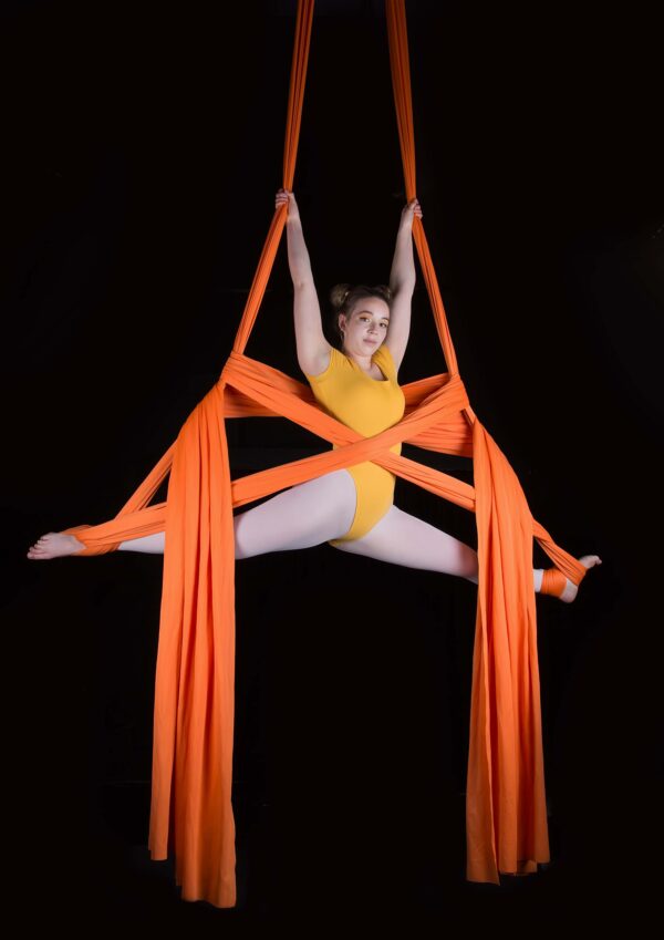 A circus performer, dressed in a yellow leotard, is suspended in orange ribbonss. The background is pure black.