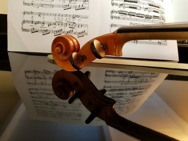 The headstock of a violin sits on a reflective surface. Sheet music stands in the background.